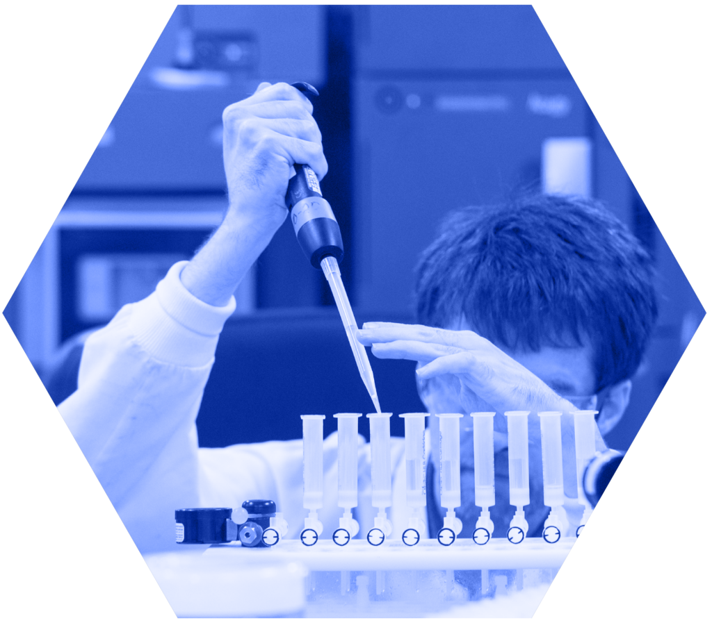 A laboratory scientist in a white coat uses a pipette to carefully dispense liquid into a row of test tubes or sample vials arranged in a rack, viewed from a close angle. The image is tinted in blue and cropped into a hexagonal shape against a black background.