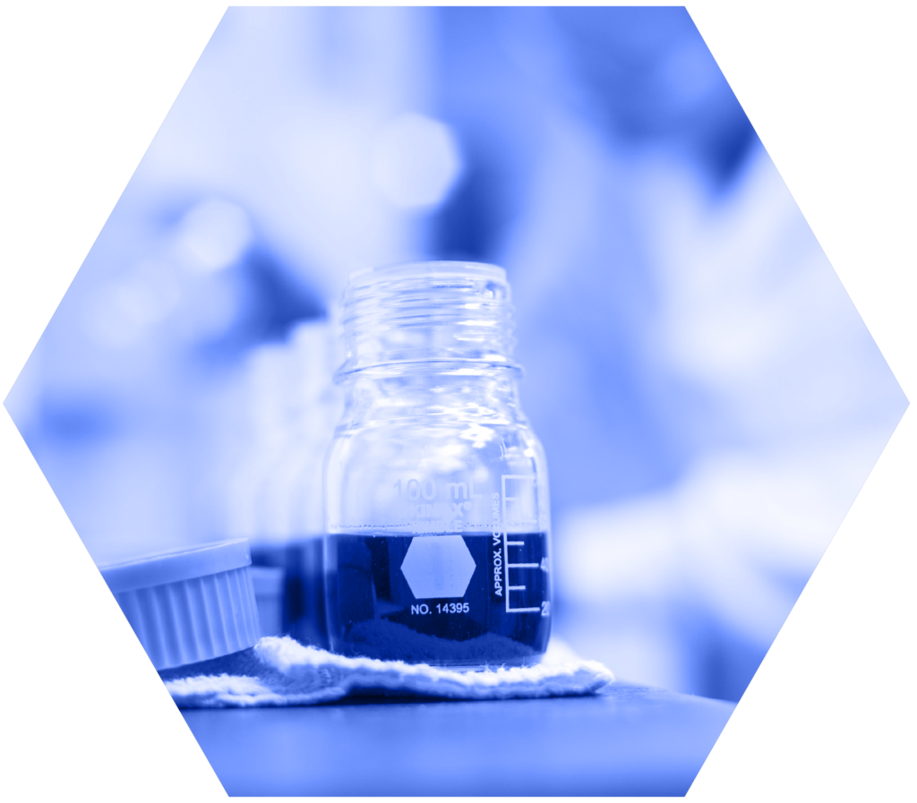 A close-up of a 100 mL glass laboratory bottle containing a dark liquid, resting on a white cloth in a laboratory setting. A white-capped container is partially visible to the left. The image has a blue tint, a soft bokeh background, and is cropped into a hexagonal shape against a black background.