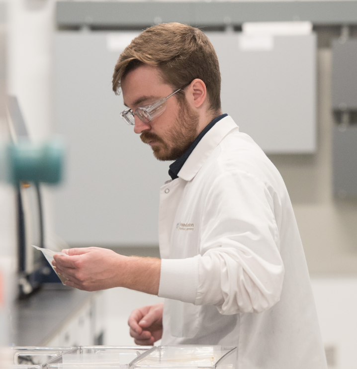 A male laboratory technician wearing a white lab coat and safety glasses examines a small document or label while standing at a lab bench, with equipment visible in the background.